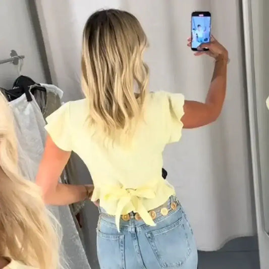Back view of woman wearing a yellow short sleeve blouse and pant set with a tied waist posing for a mirror selfie