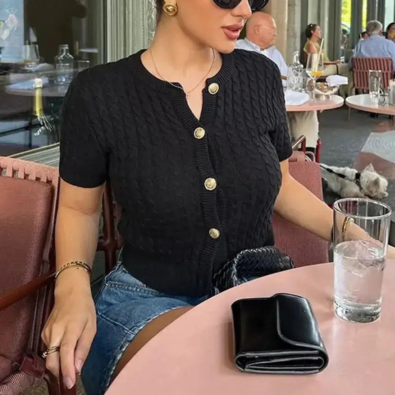 Black cropped sweater cardigan with gold buttons worn by woman sitting at table with glass of water