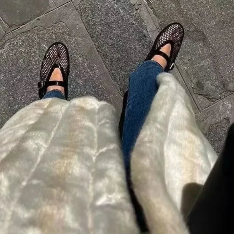 Black mesh fashion sandals worn with blue jeans and white textured coat on stone pavement