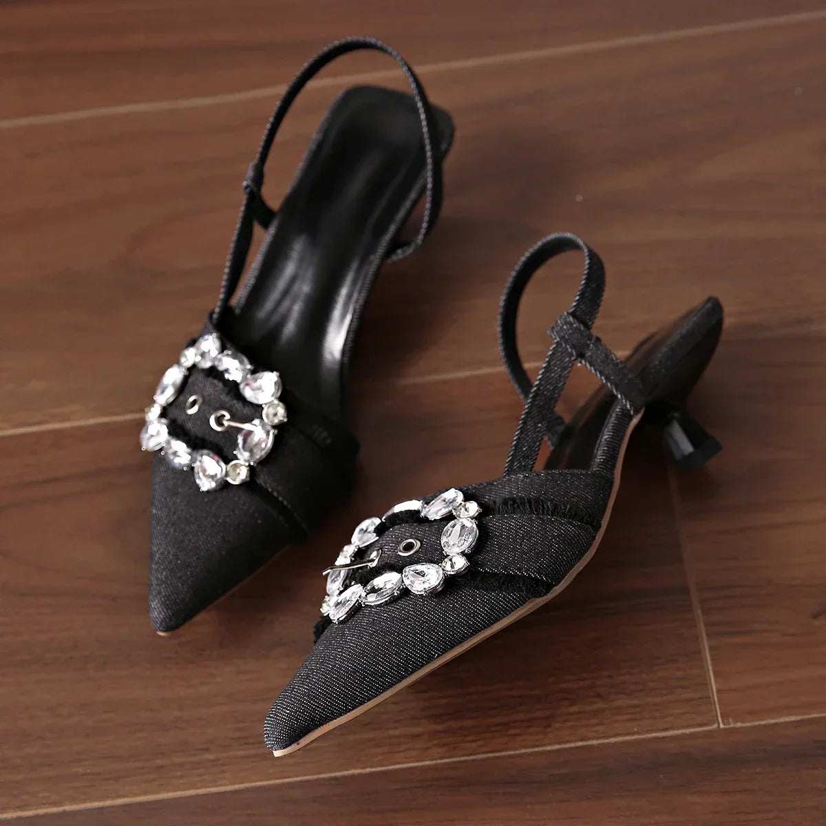 Black pointed toe stiletto sandals with sparkling crystal buckle and slingback strap on wooden floor