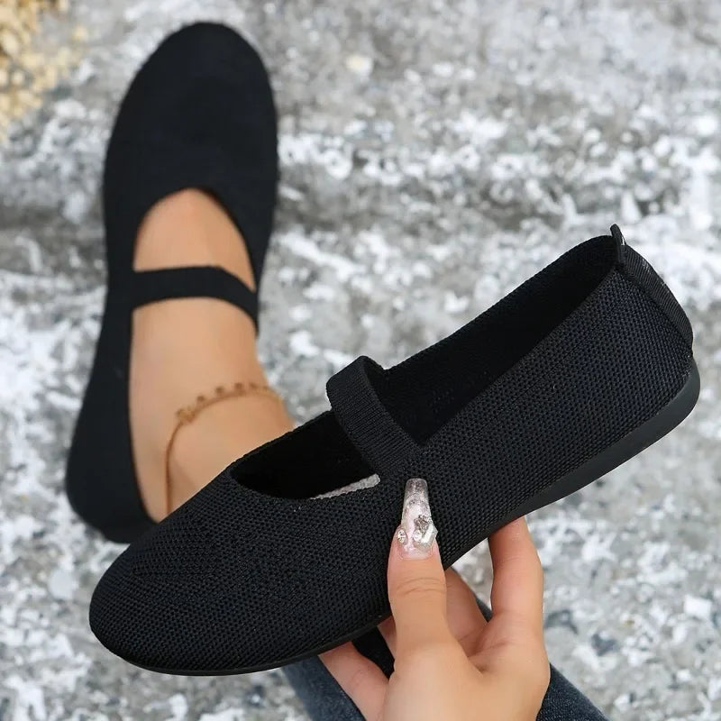 Black comfortable slip-on shoes with strap detail held by hand and worn on foot on textured ground