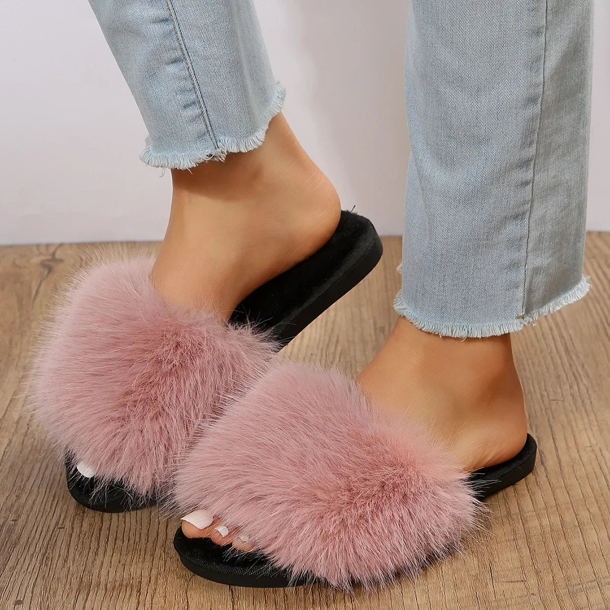 Pink fuzzy flat slide slippers with black soles worn with light blue jeans on wooden floor