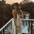 Woman wearing a sleeveless satin formal dress with spaghetti straps standing on a balcony at sunset
