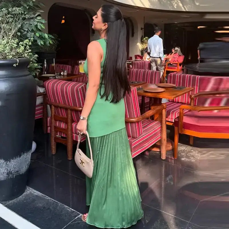 Woman wearing sleeveless green maxi dress with pleated skirt holding a beige handbag indoors